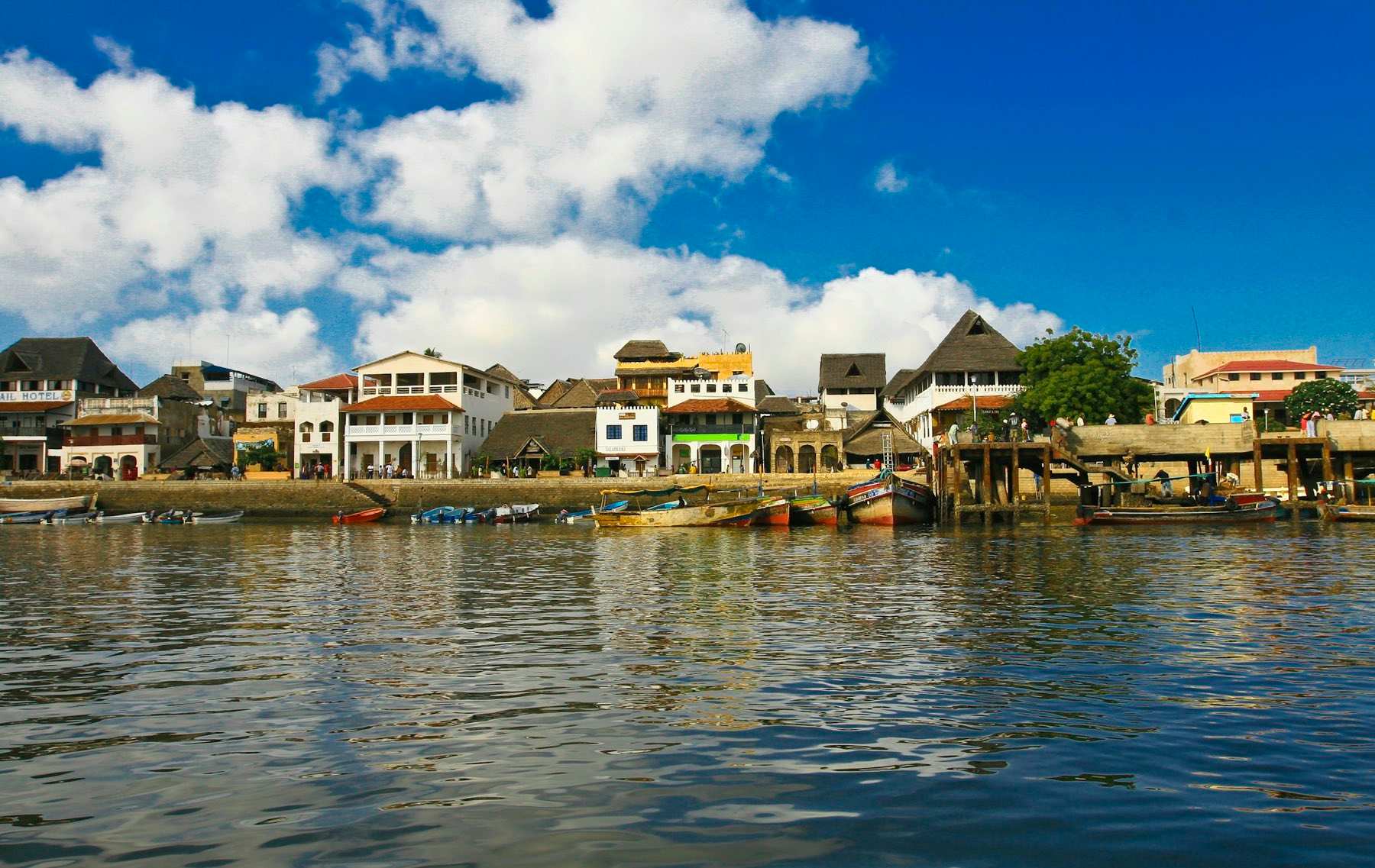 Stone houses and makuti roofs along the Lamu waterfront, fishing boats moored at the jetty