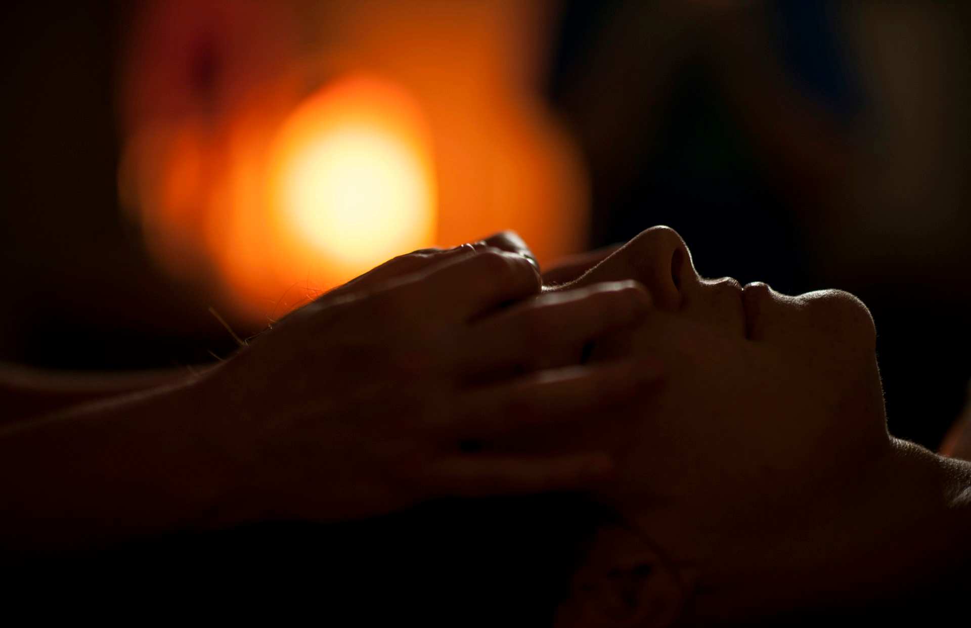 Hands working across a person's face during a massage, a warm lamp glowing in the dark behind