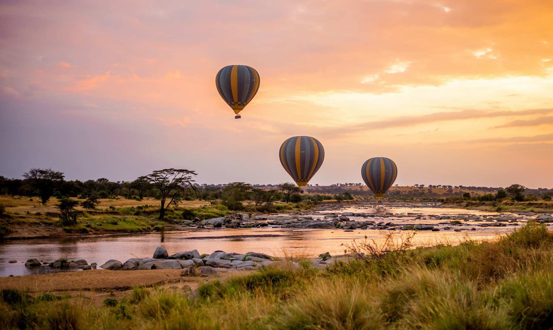 Three hot-air balloons drift over a rocky river in the Serengeti at dawn