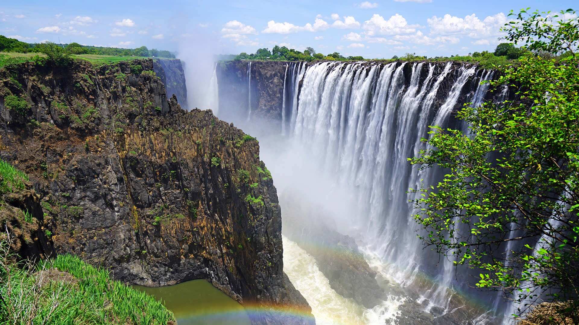 Victoria Falls dropping in strands over a dark basalt cliff, spray rising from the gorge and a rainbow in the chasm below